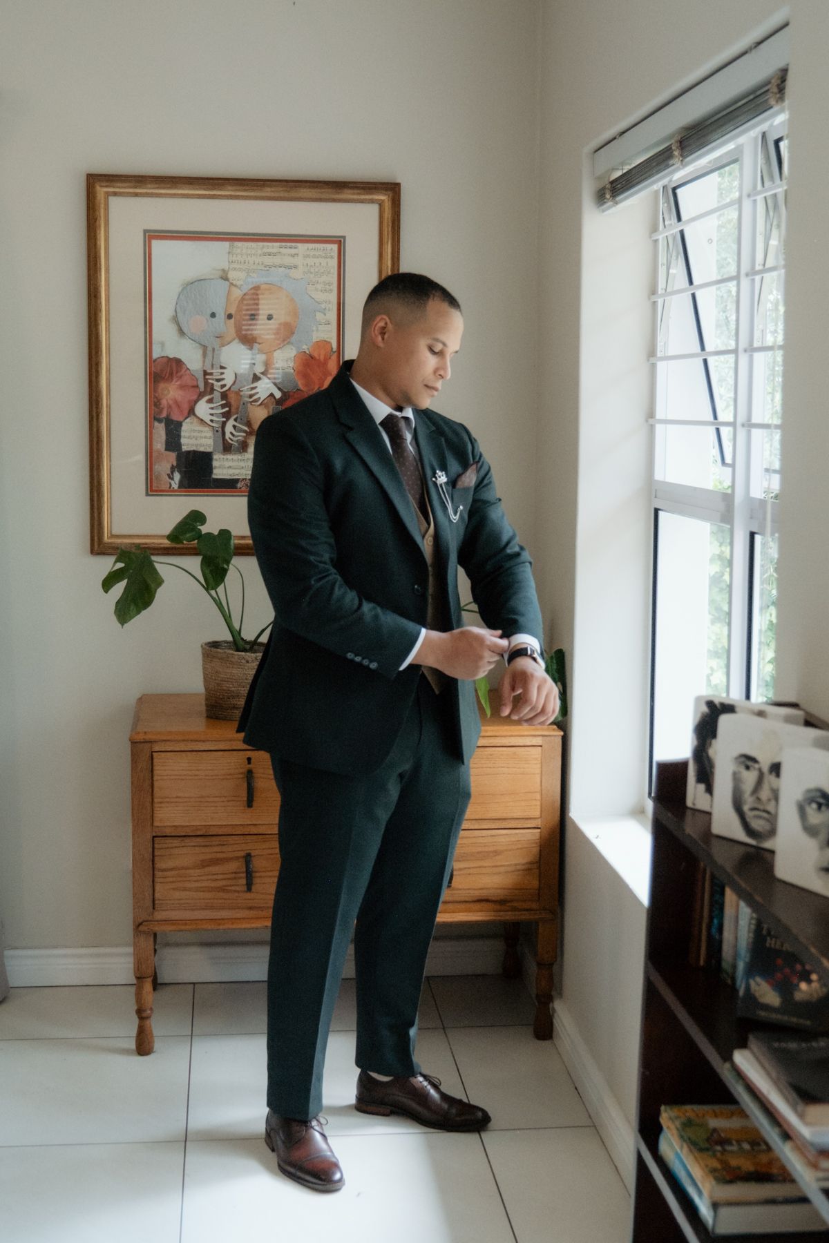Groom in dark green suit checking his watch while standing by a window before wedding.