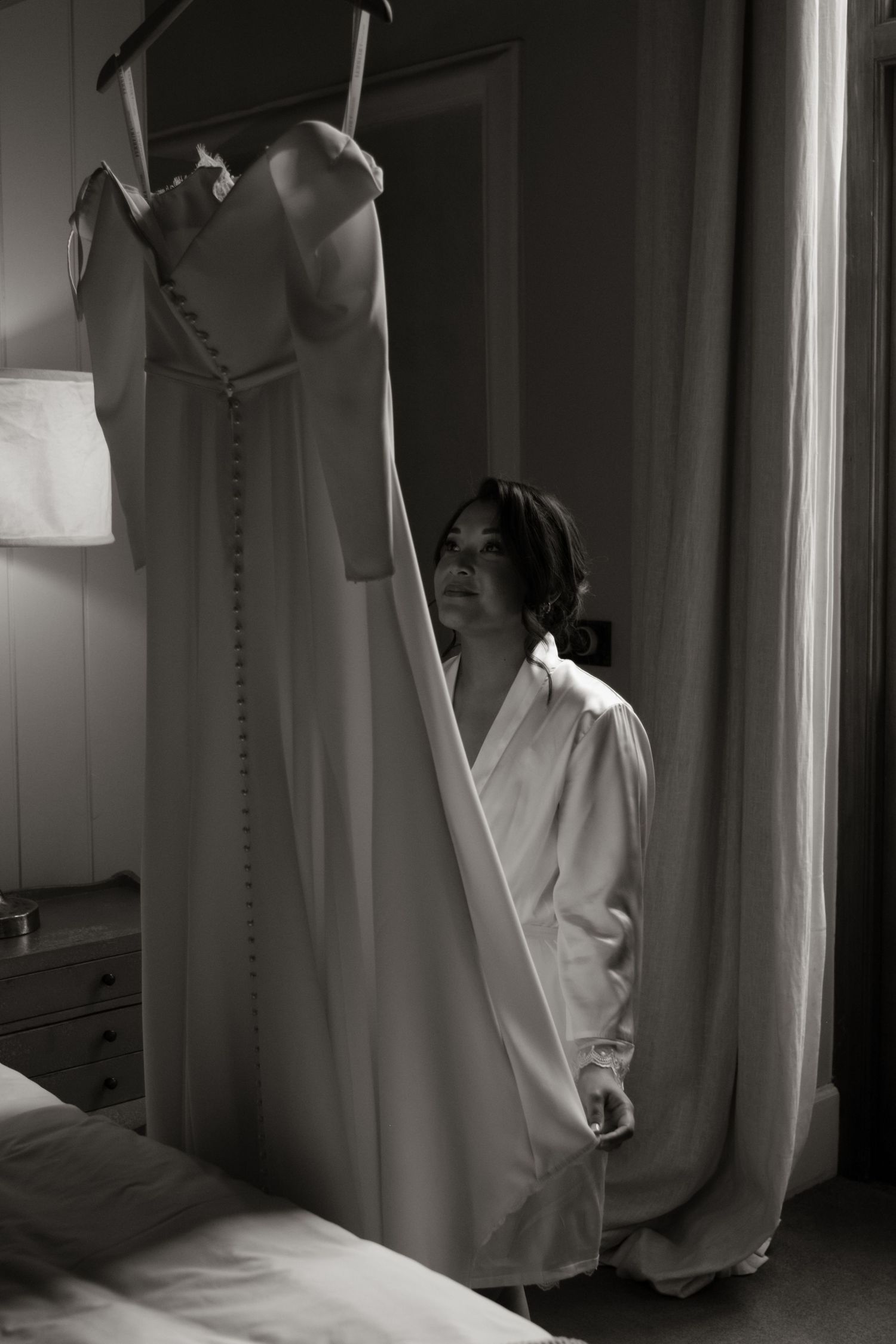 Bride in white robe admiring her wedding dress hanging in a softly lit hotel room, black and white photo.