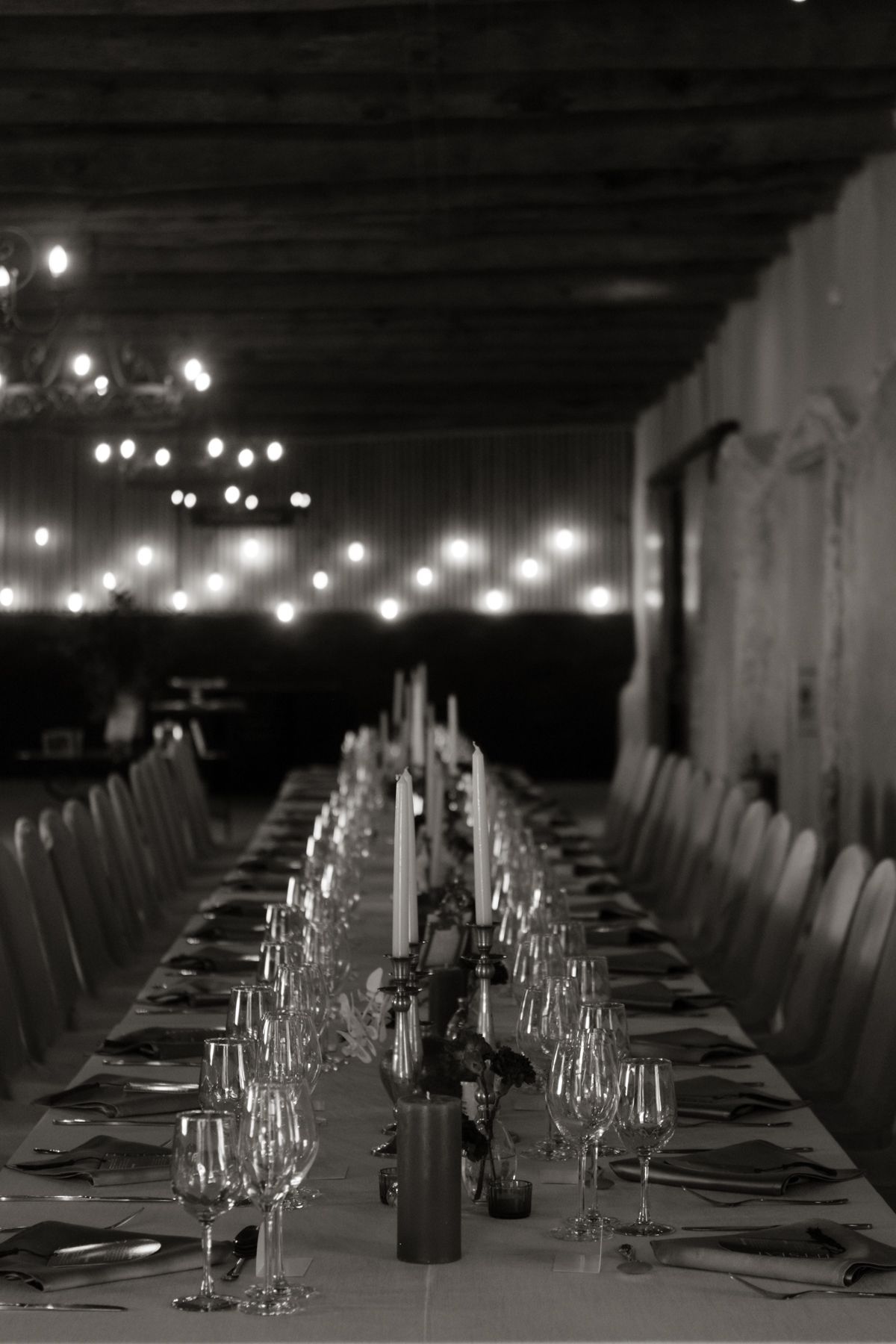 Dramatic black and white photo of long wedding banquet table lit by candles and string lights.