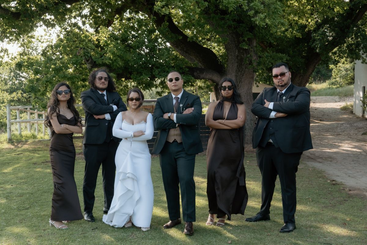 Wedding party of six posing with crossed arms in sunglasses under a large tree at an outdoor ceremony.