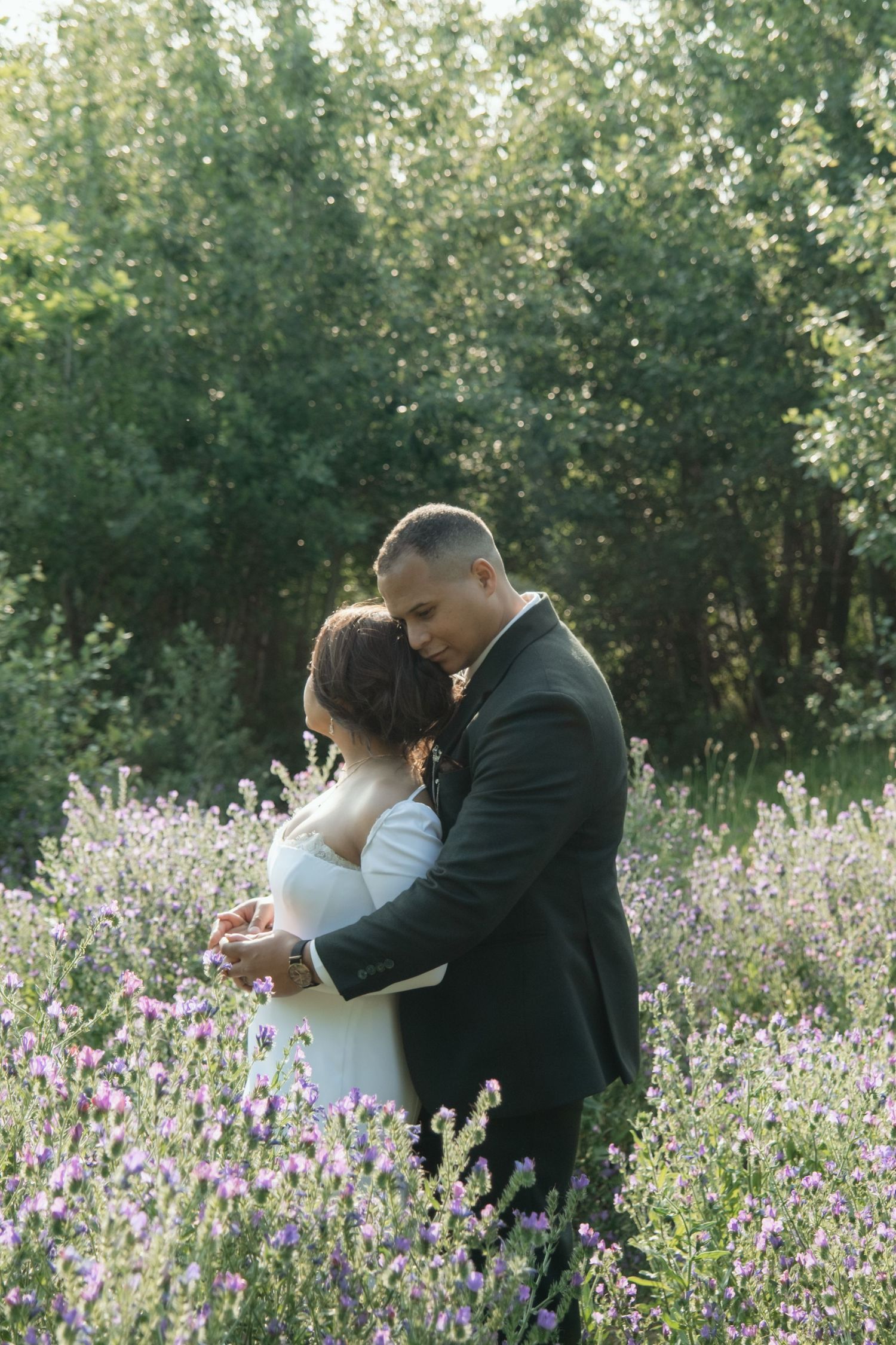 Newlywed couple embraces tenderly in a lavender field surrounded by lush green trees at golden hour.
