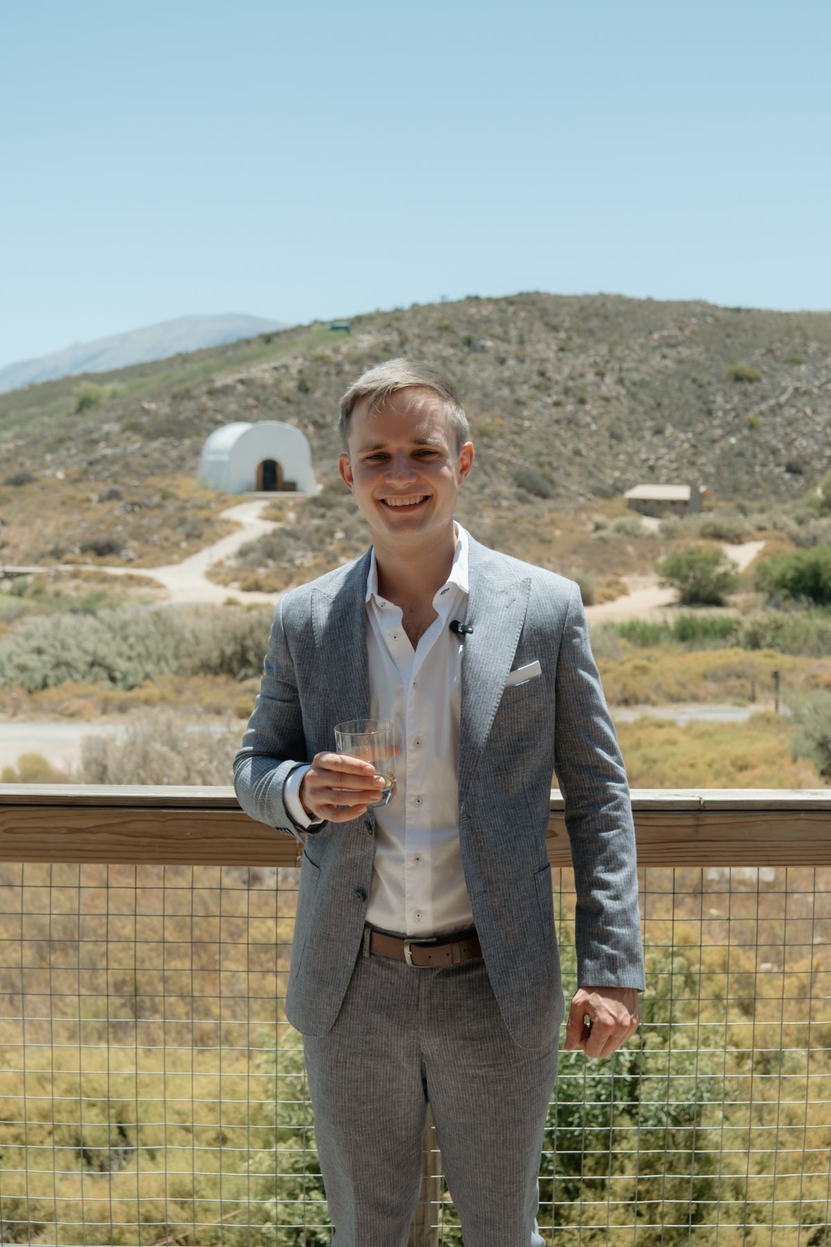 Smiling groom in grey suit holding champagne glass on outdoor deck with desert landscape and hills behind him.