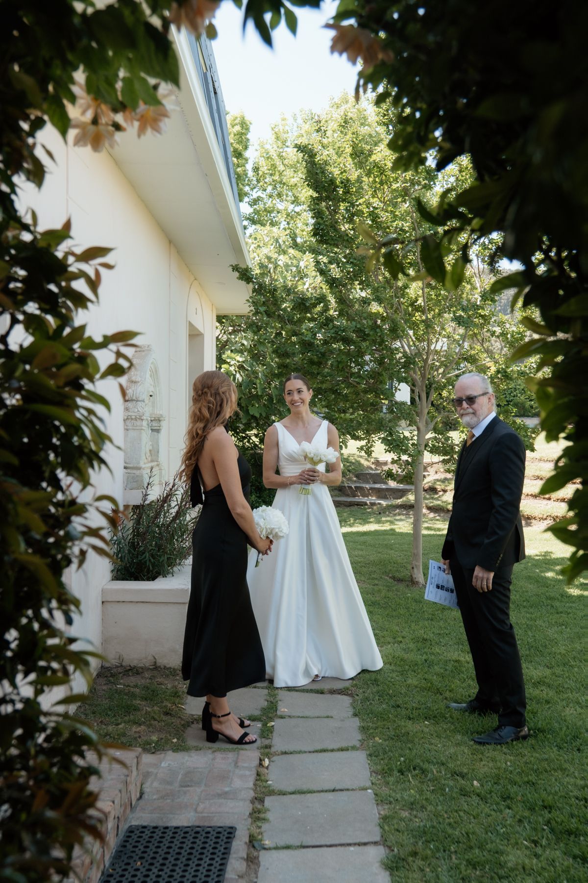 Bride in white gown holds bouquet while chatting with guests outside white building on wedding day.
