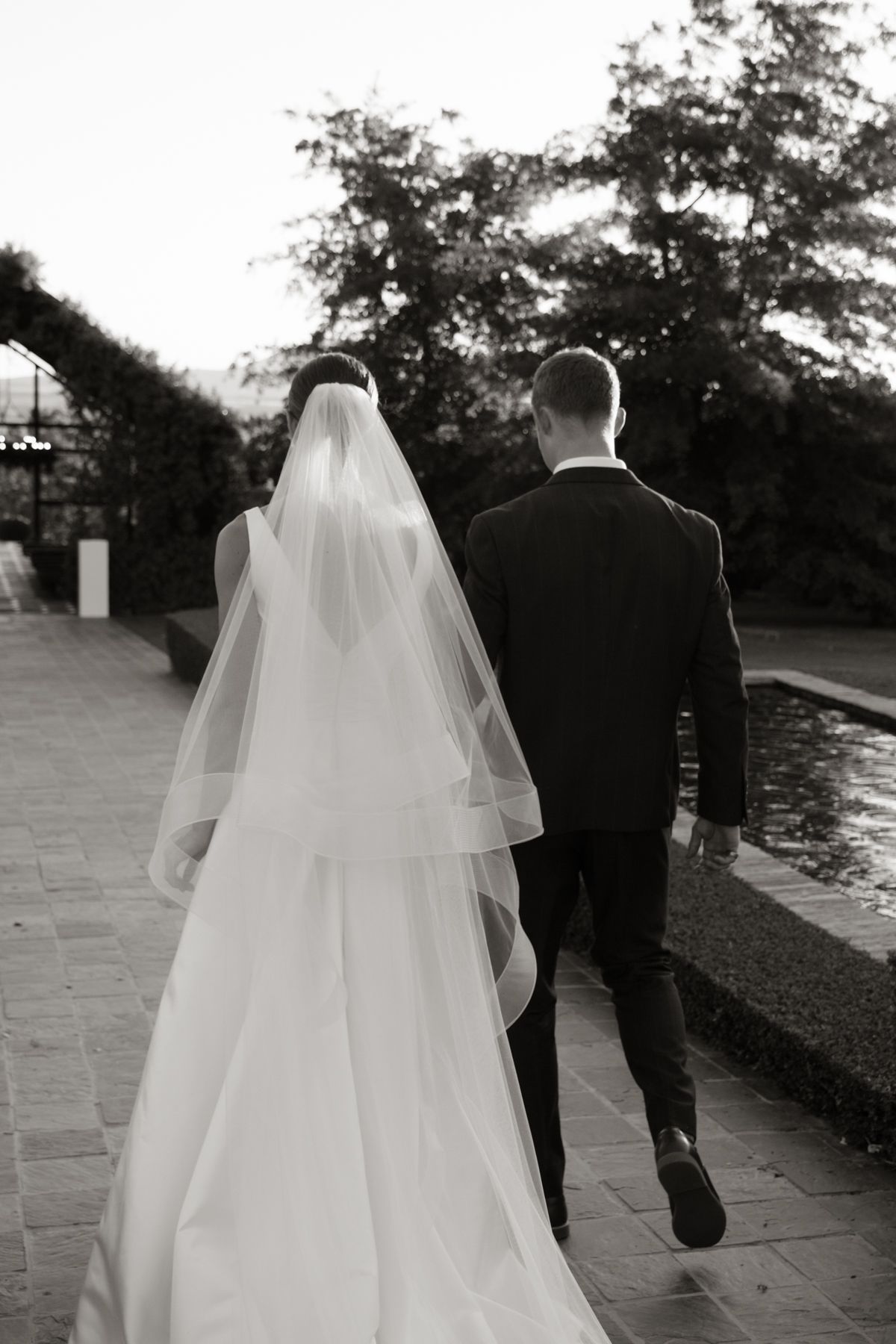 Black and white photo of bride and groom walking hand in hand along garden path by pond.