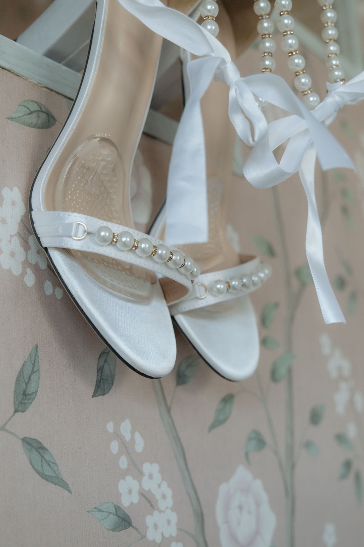 Elegant white bridal sandals with pearl embellishments and ribbon ties displayed beside a pearl necklace on floral background.