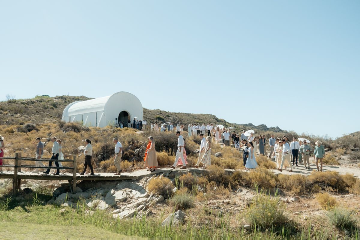 Crowd of wedding guests gathered outside a white dome chapel surrounded by dry shrubland on a clear sunny day.