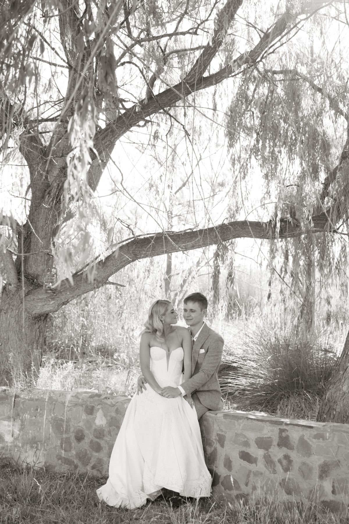 Bride and groom embracing by stone wall under a weeping tree, black and white photo.