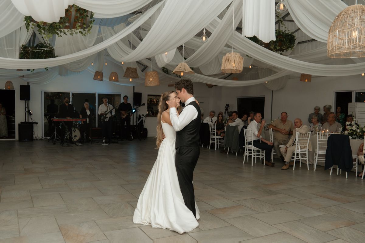 Bride and groom sharing first dance under white draped ceiling with wedding guests seated in background.