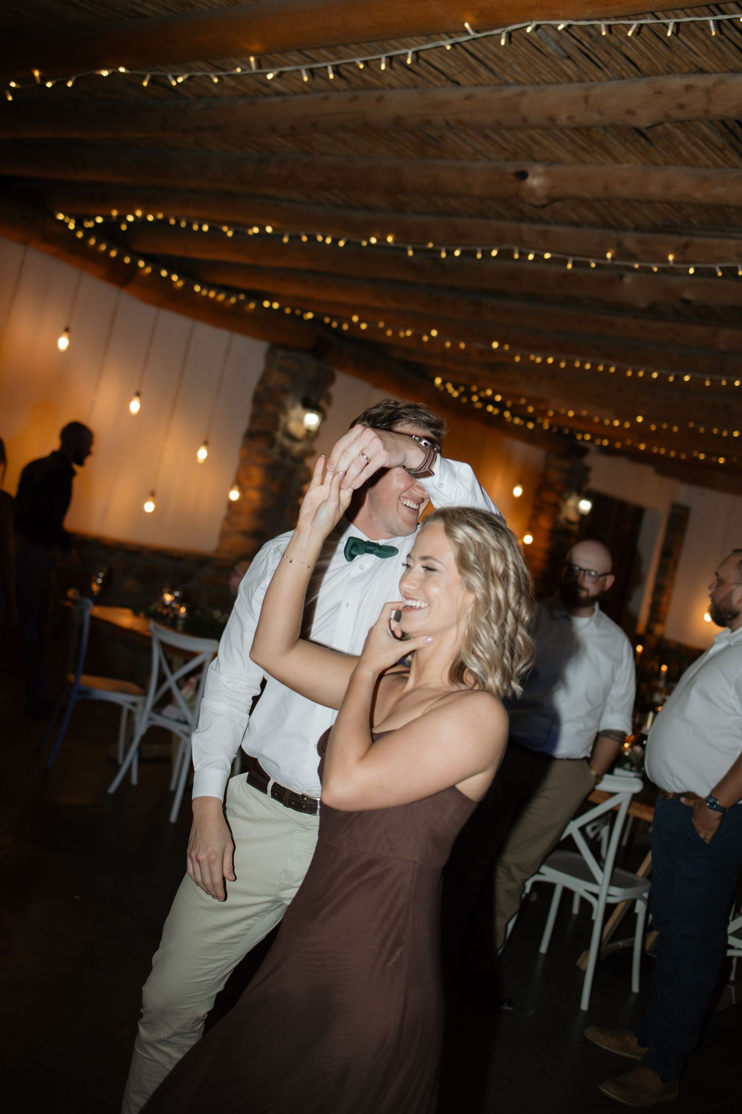 Couple dancing joyfully under warm string lights at rustic indoor wedding reception venue.