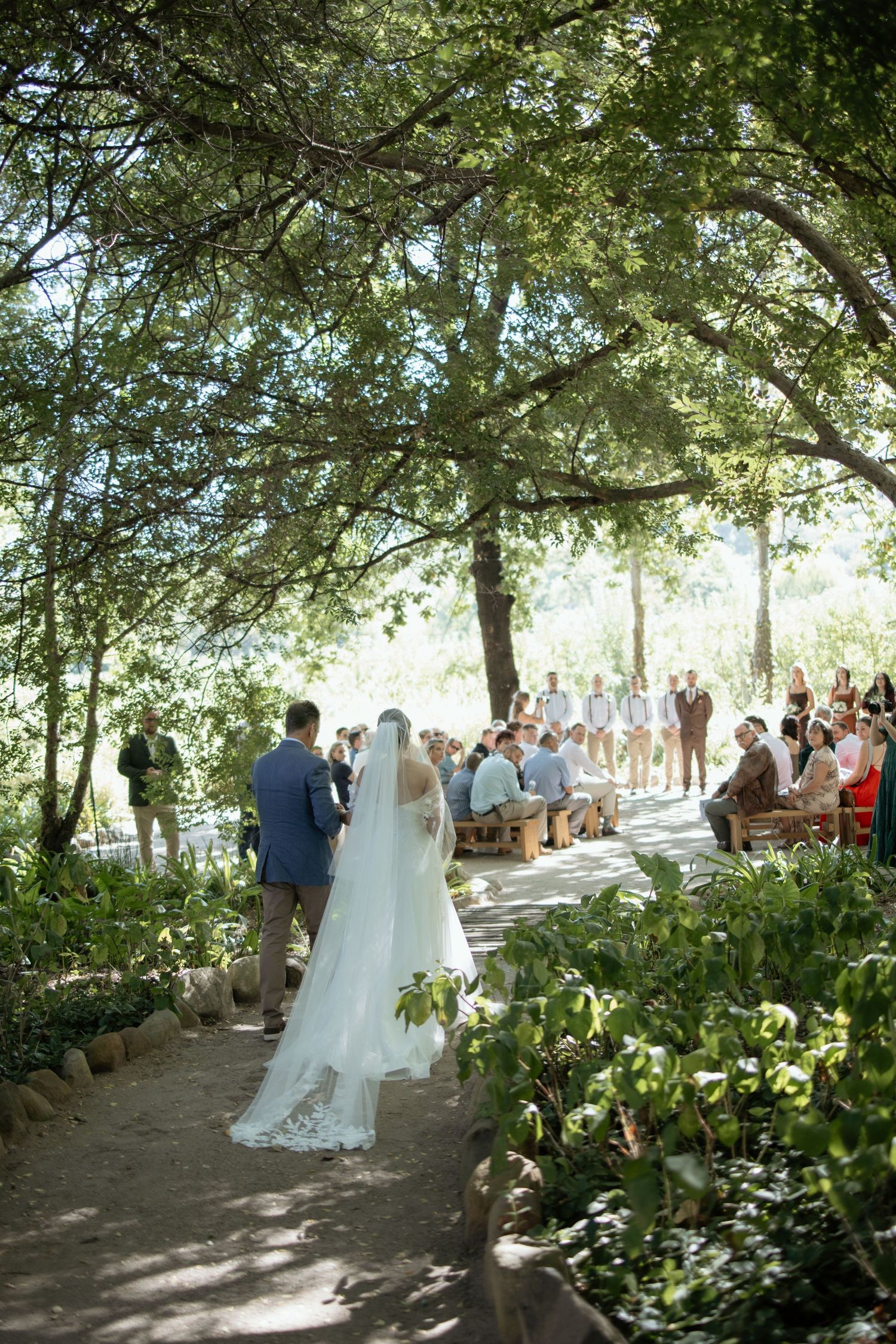 Bride walks down an outdoor garden wedding aisle lined with lush green trees and seated guests.
