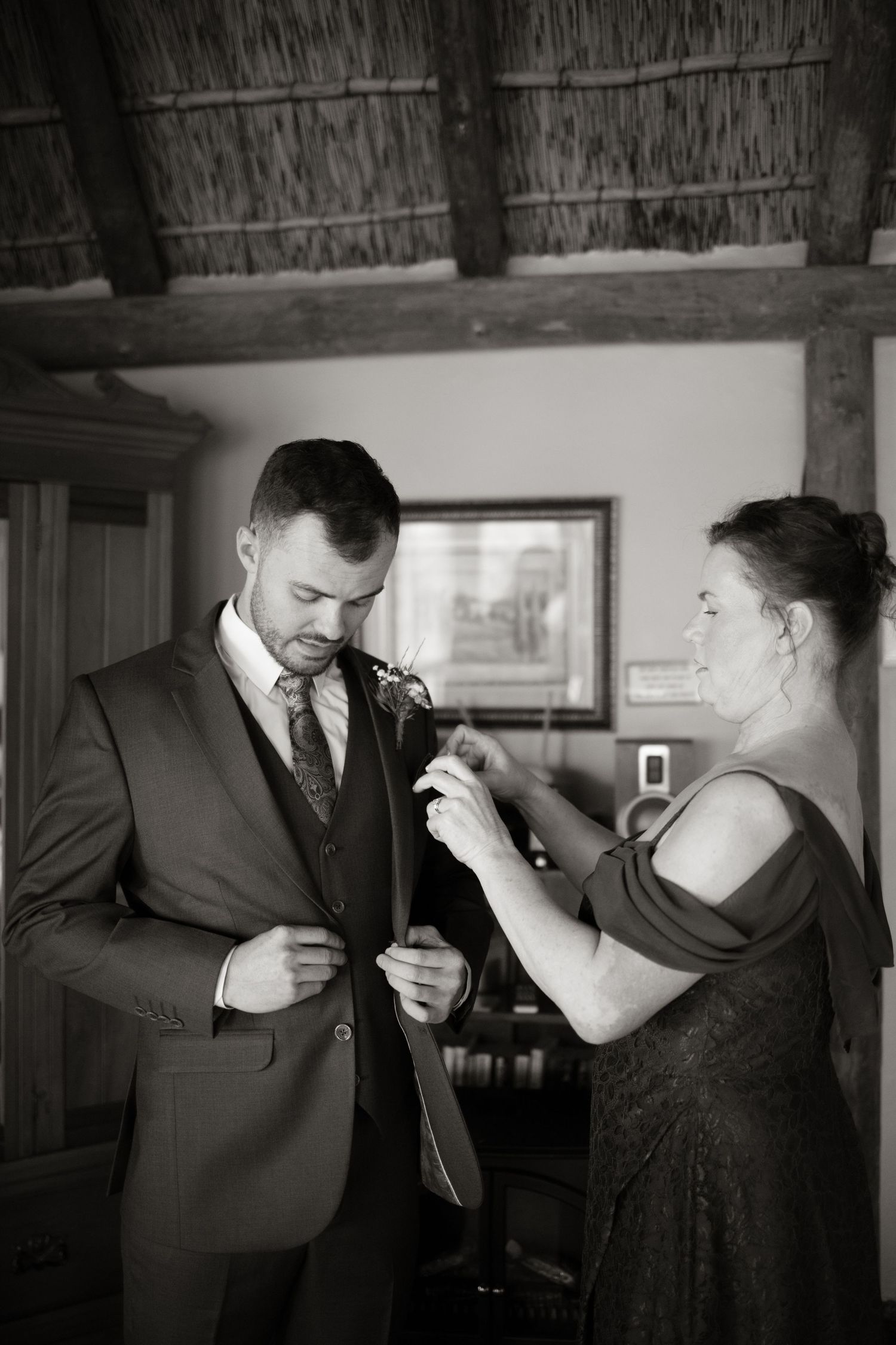 Black and white photo of a woman helping a groom pin a boutonniere on his suit jacket before his wedding.