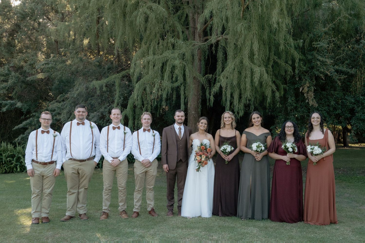 Wedding party poses under large evergreen tree with groomsmen in suspenders and bridesmaids in earth tones.