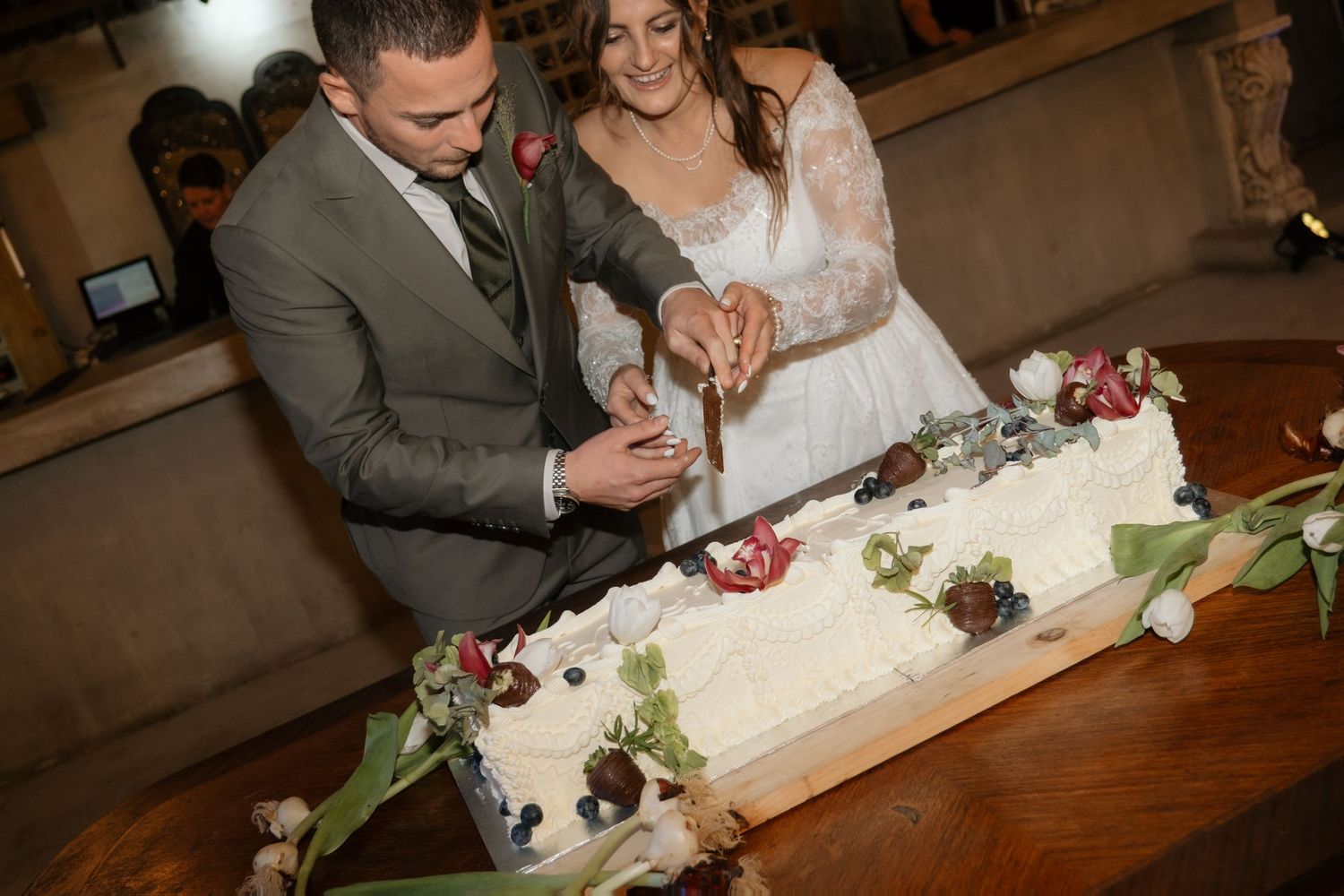 Bride and groom cutting long white wedding cake decorated with flowers and berries.