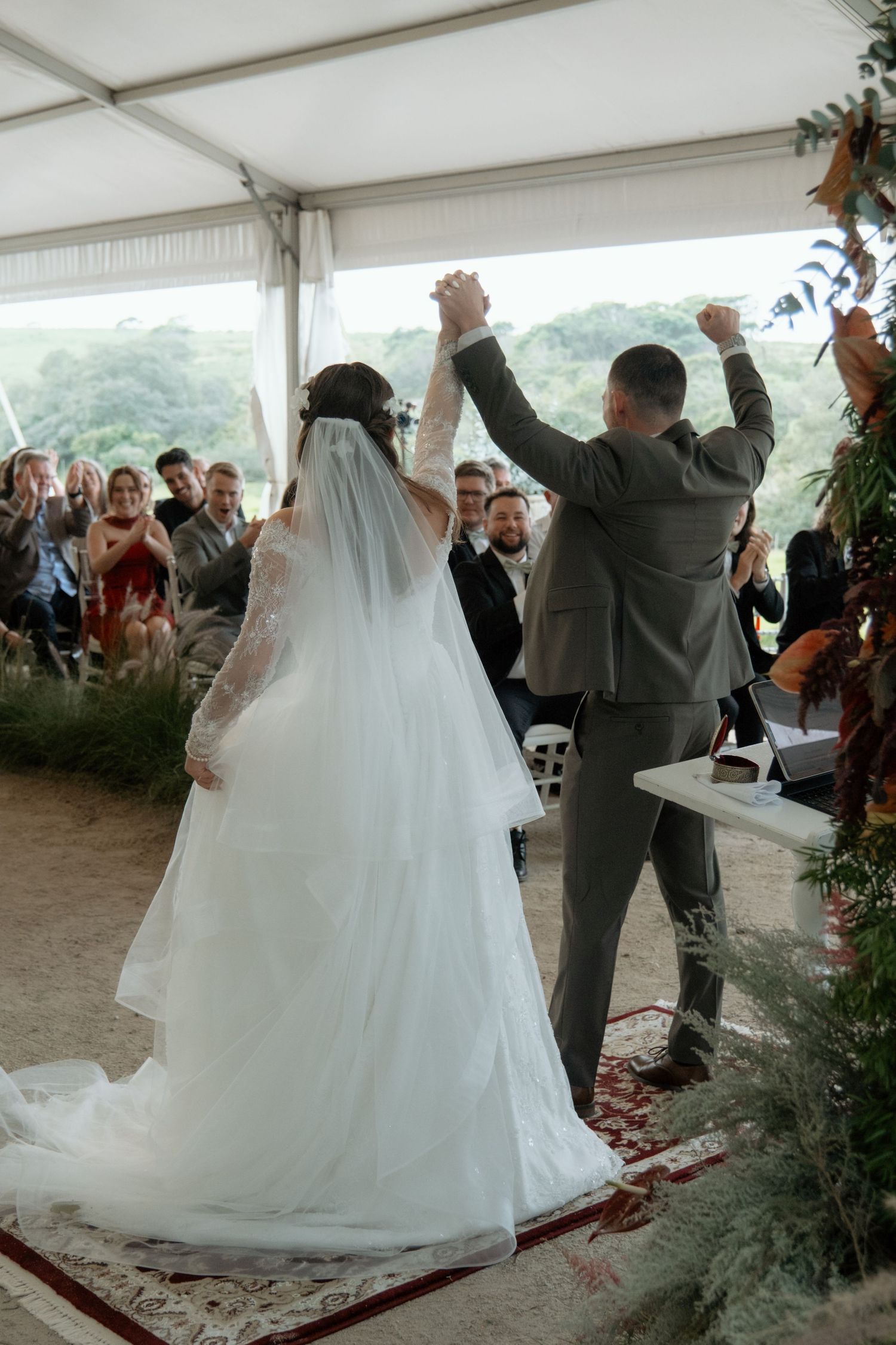 Newlyweds raise joined hands in celebration at outdoor wedding ceremony with guests applauding.
