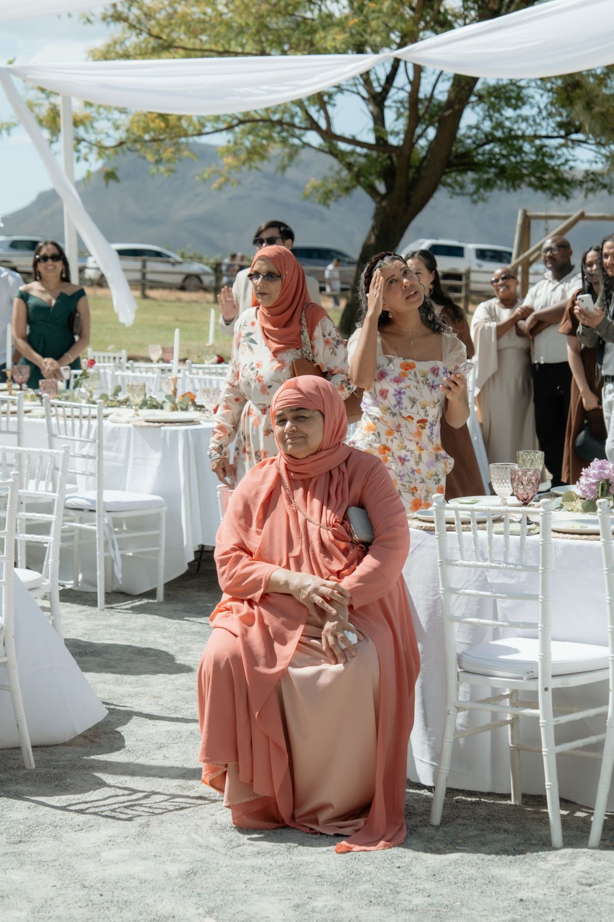 Muslim wedding guests in pink hijabs watch an outdoor ceremony at elegantly decorated white tables.