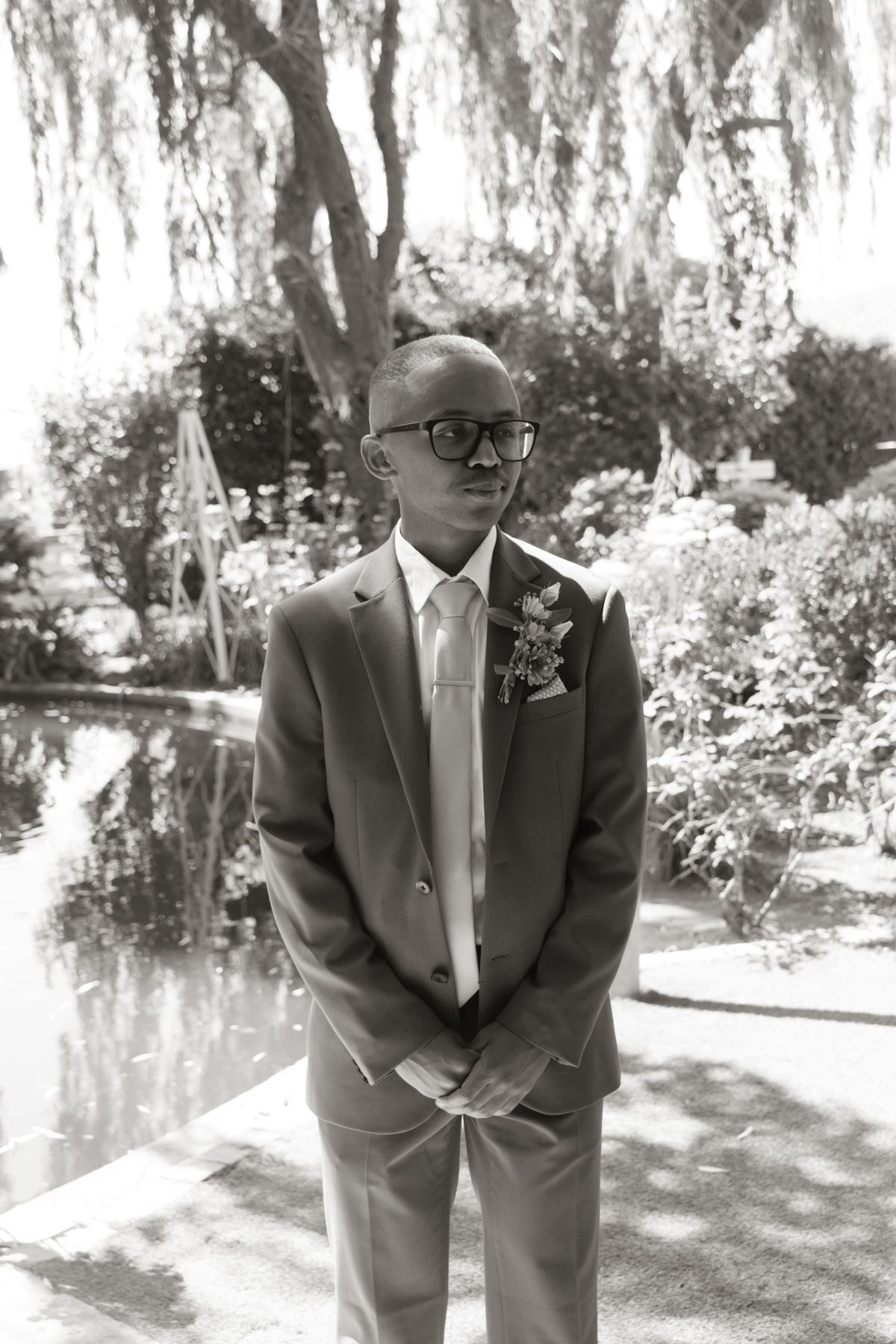 Groom in brown suit with boutonniere stands in lush garden, black and white wedding portrait.