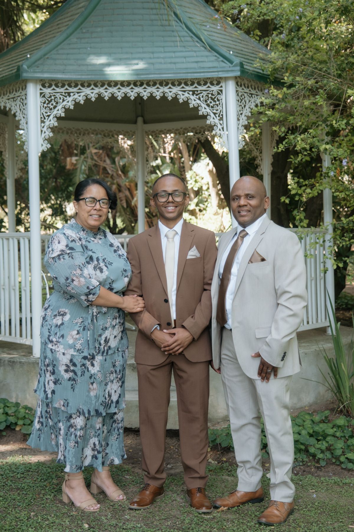 Groom in brown suit poses with parents in front of white decorative garden gazebo.