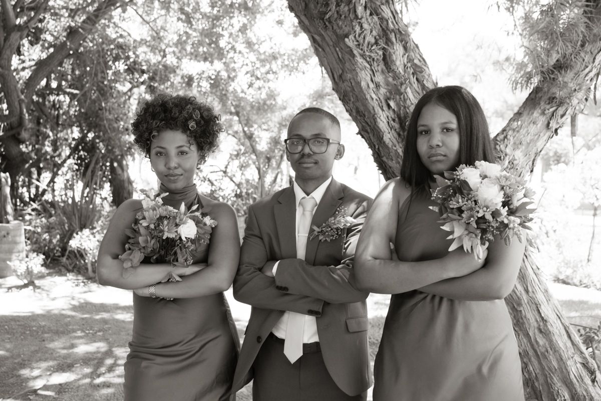 Black and white photo of groom in suit posing with two bridesmaids holding bouquets in a playful serious pose.