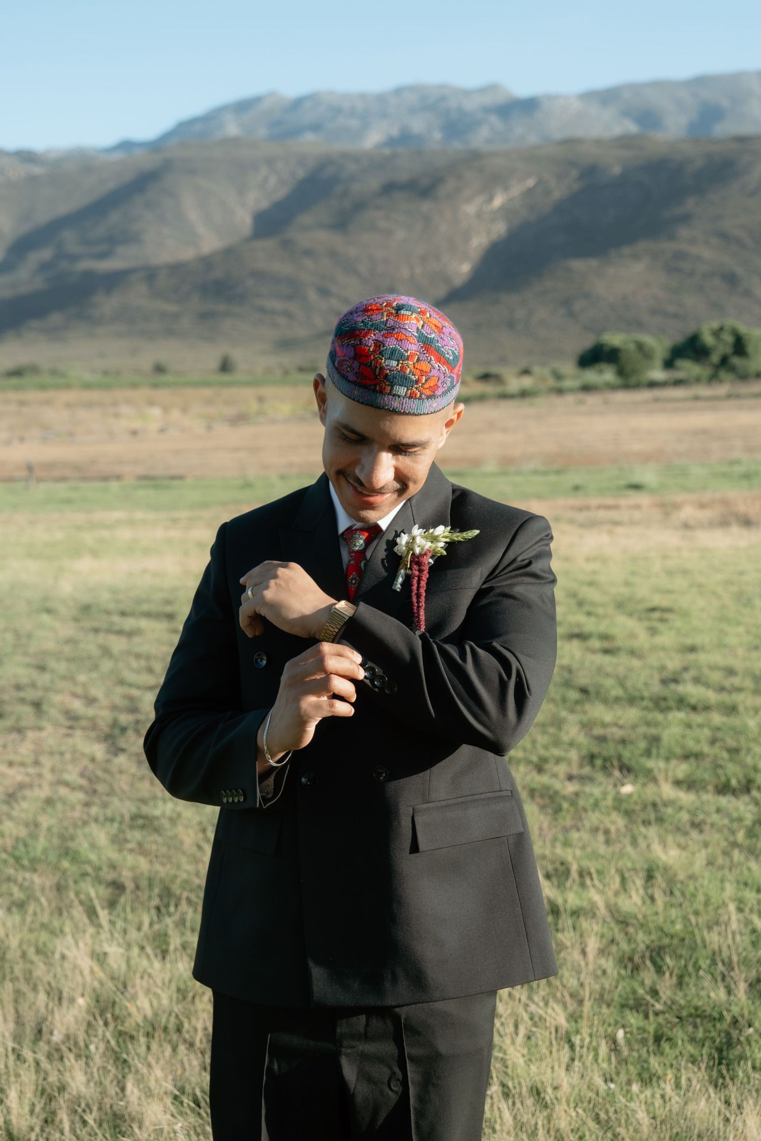 Groom in black suit and colorful kufi cap adjusting cufflinks outdoors with mountain backdrop.