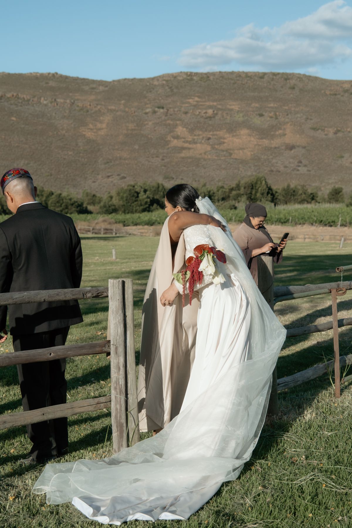 Bride in white gown with long veil walking through wooden fence gate in mountain landscape.