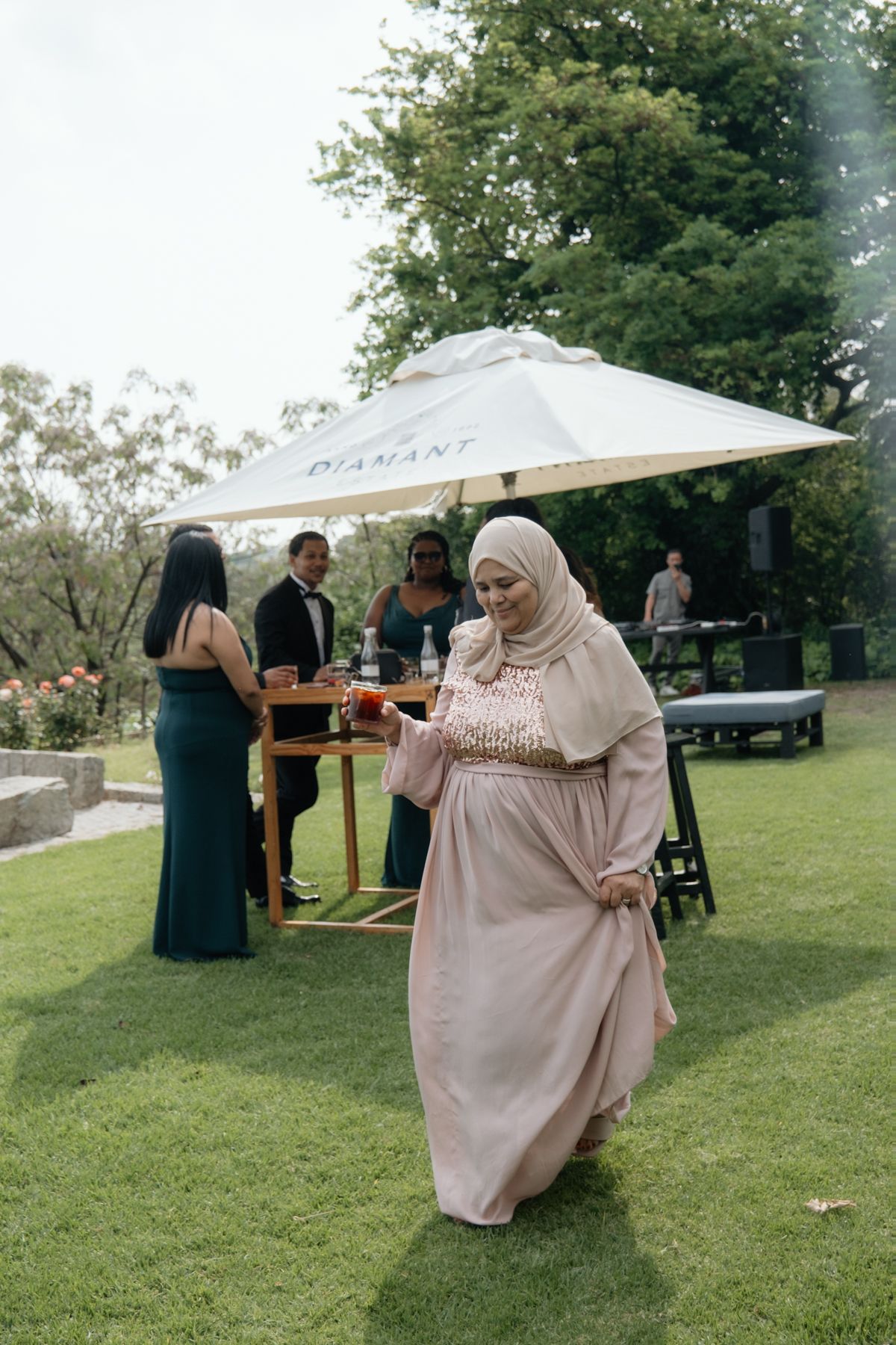 A woman in a hijab and blush gown walks across a lawn at an outdoor wedding reception.