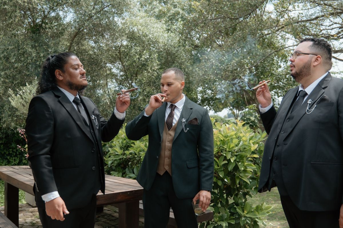 Three groomsmen in dark suits enjoy cigars together outdoors surrounded by lush green trees before wedding.