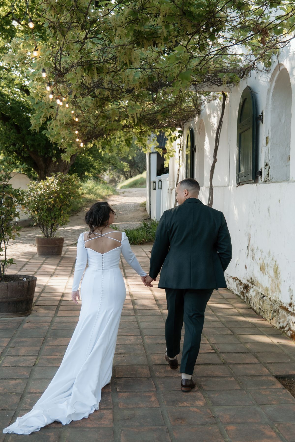 Bride and groom walking hand in hand along brick pathway beside white building draped with lush green vines.