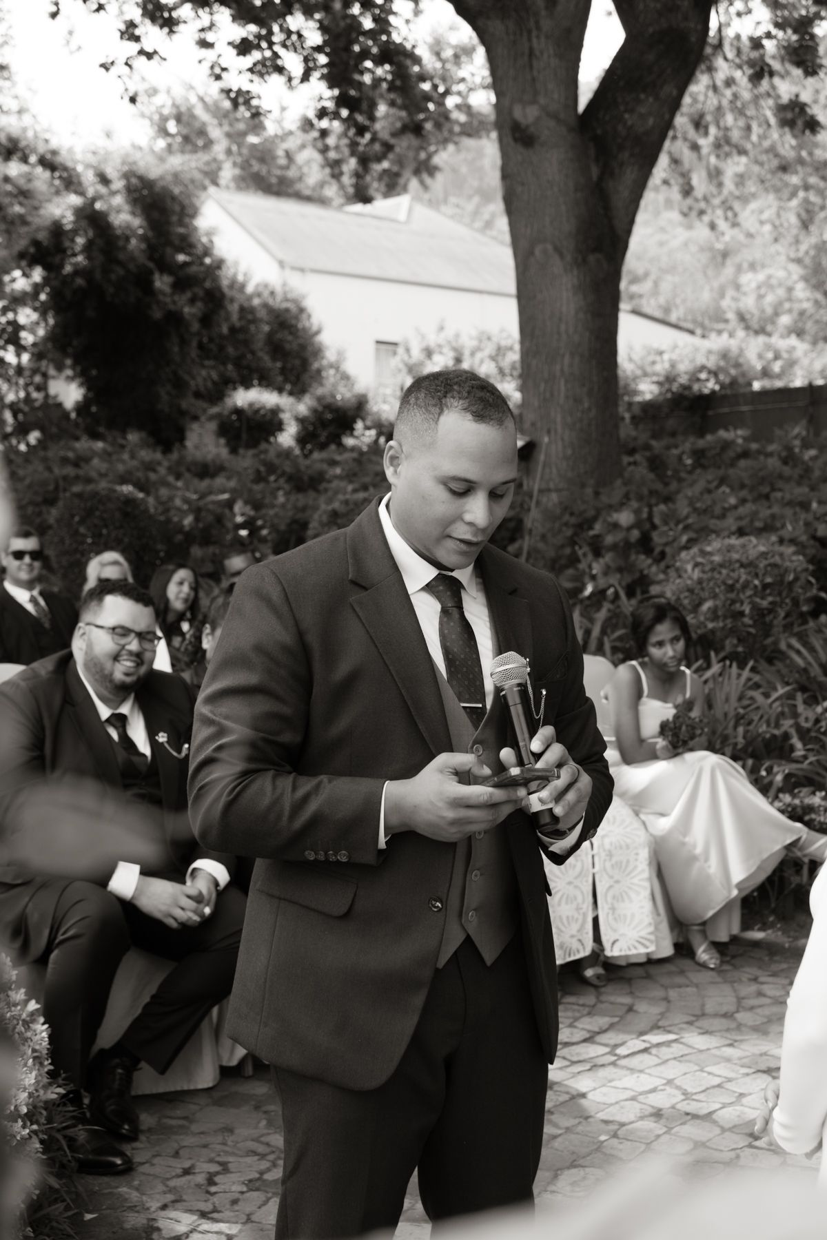 Groom in a dark suit holding a bottle during an outdoor wedding ceremony in black and white.