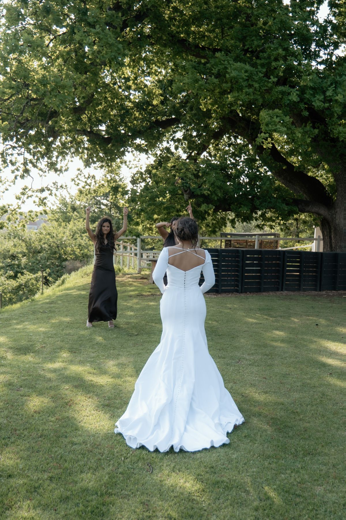 Bride in white mermaid gown poses outdoors on green lawn while photographer captures her wedding portrait under large tree.