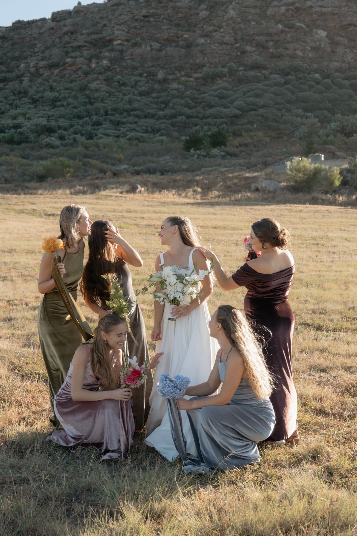 Bride with bouquet surrounded by bridesmaids in colorful gowns in rustic outdoor field setting.