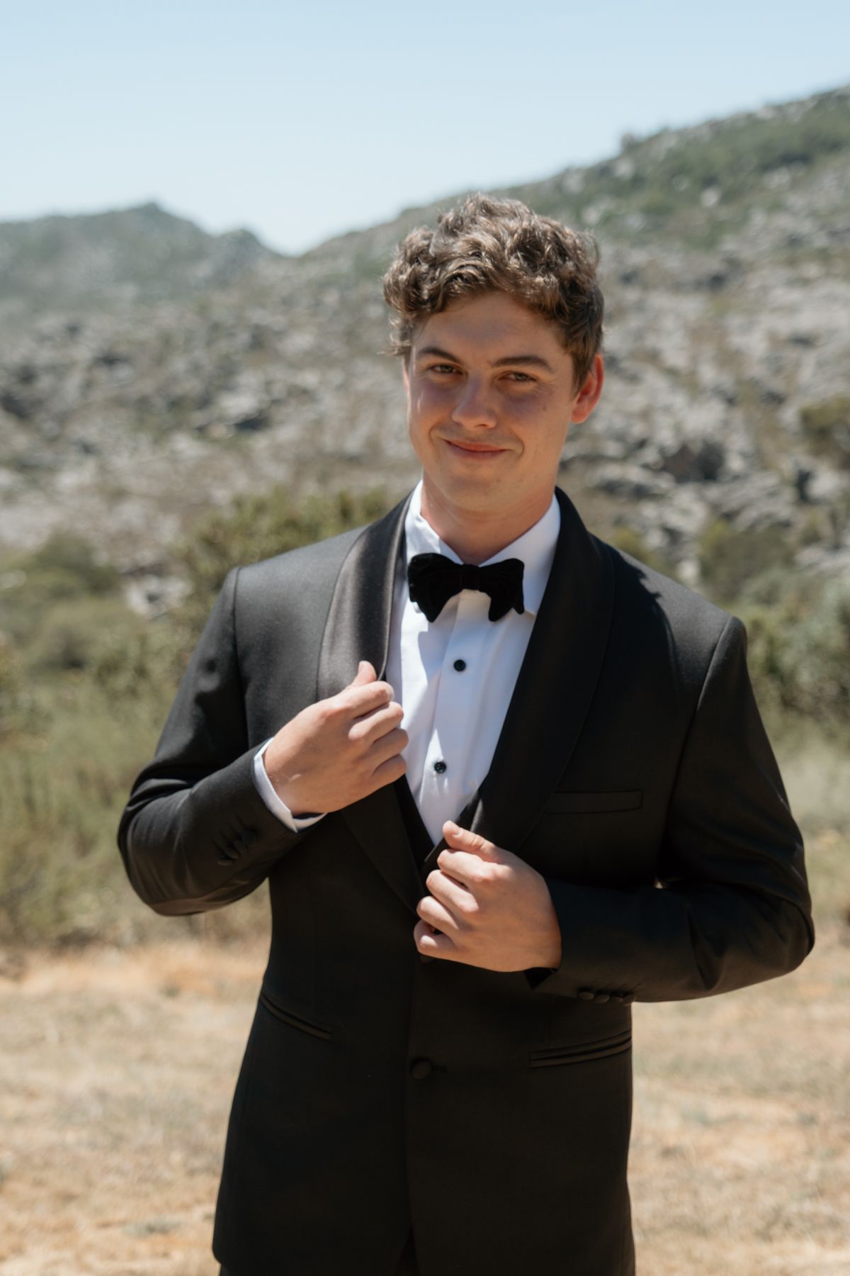 Handsome groom adjusting black tuxedo jacket with bow tie in sunny outdoor mountain setting.