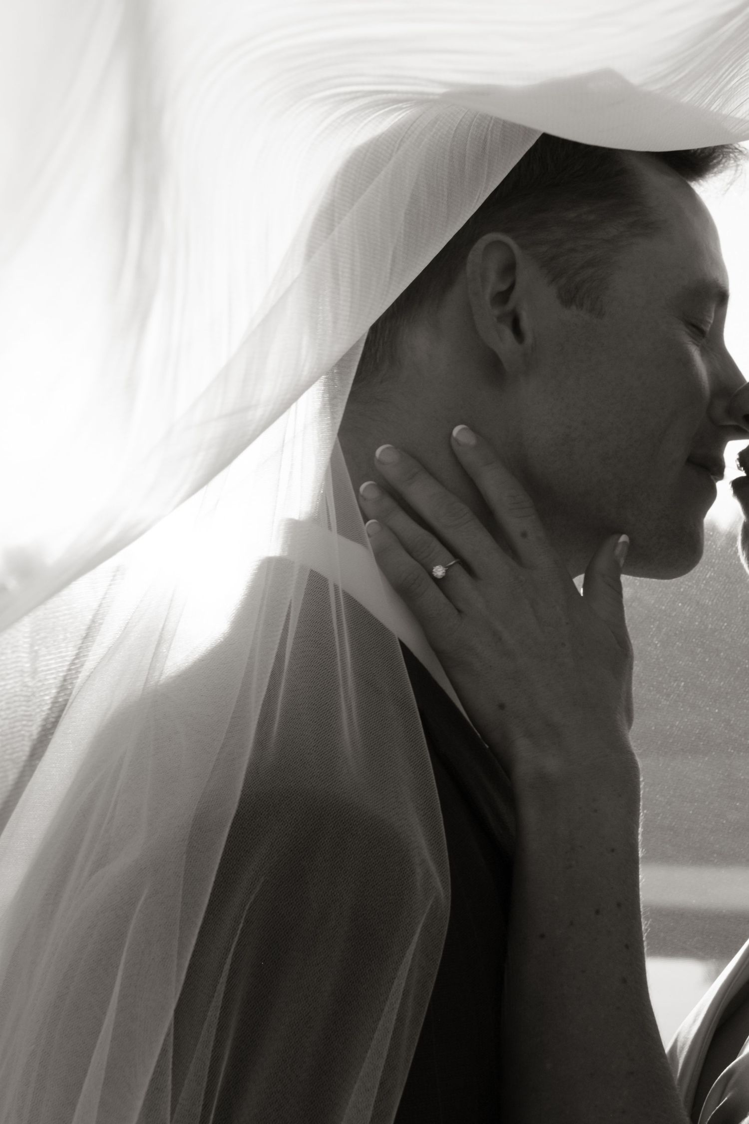 Black and white photo of bride in veil tenderly holding grooms face during intimate wedding moment.