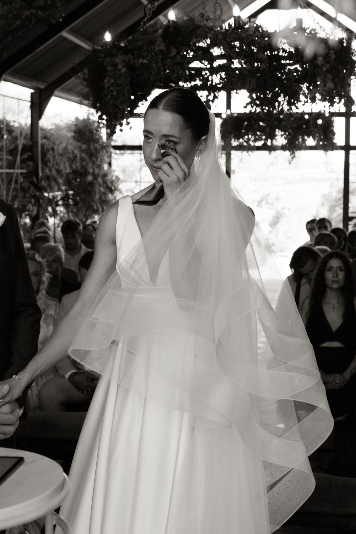 Emotional bride in white gown and veil wipes tears during wedding ceremony in black and white.
