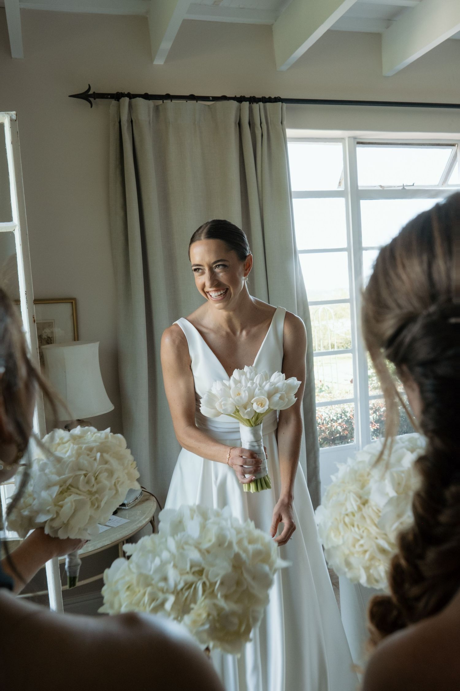 Smiling bride in white gown holds bouquet of white flowers, standing in bright room with bridesmaids nearby.