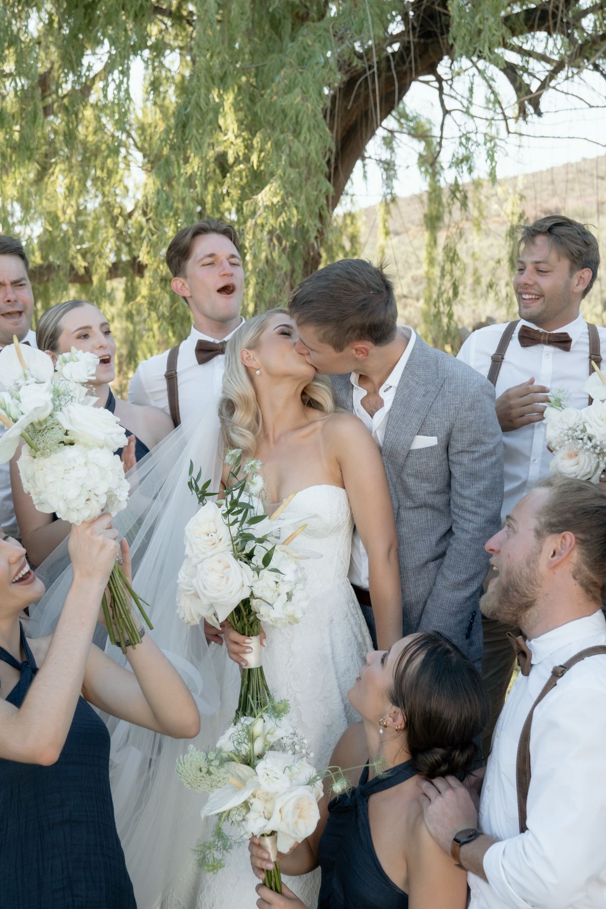 Bride and groom share a kiss surrounded by cheering wedding party under a willow tree, bridesmaids holding white floral bouquets.