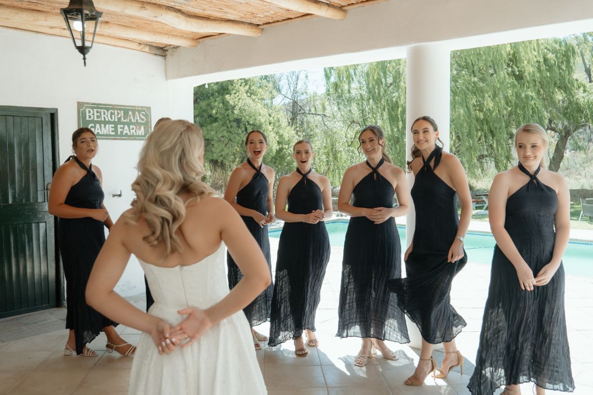 Bride in white gown reveals herself to bridesmaids in black halter dresses at a farm wedding venue.