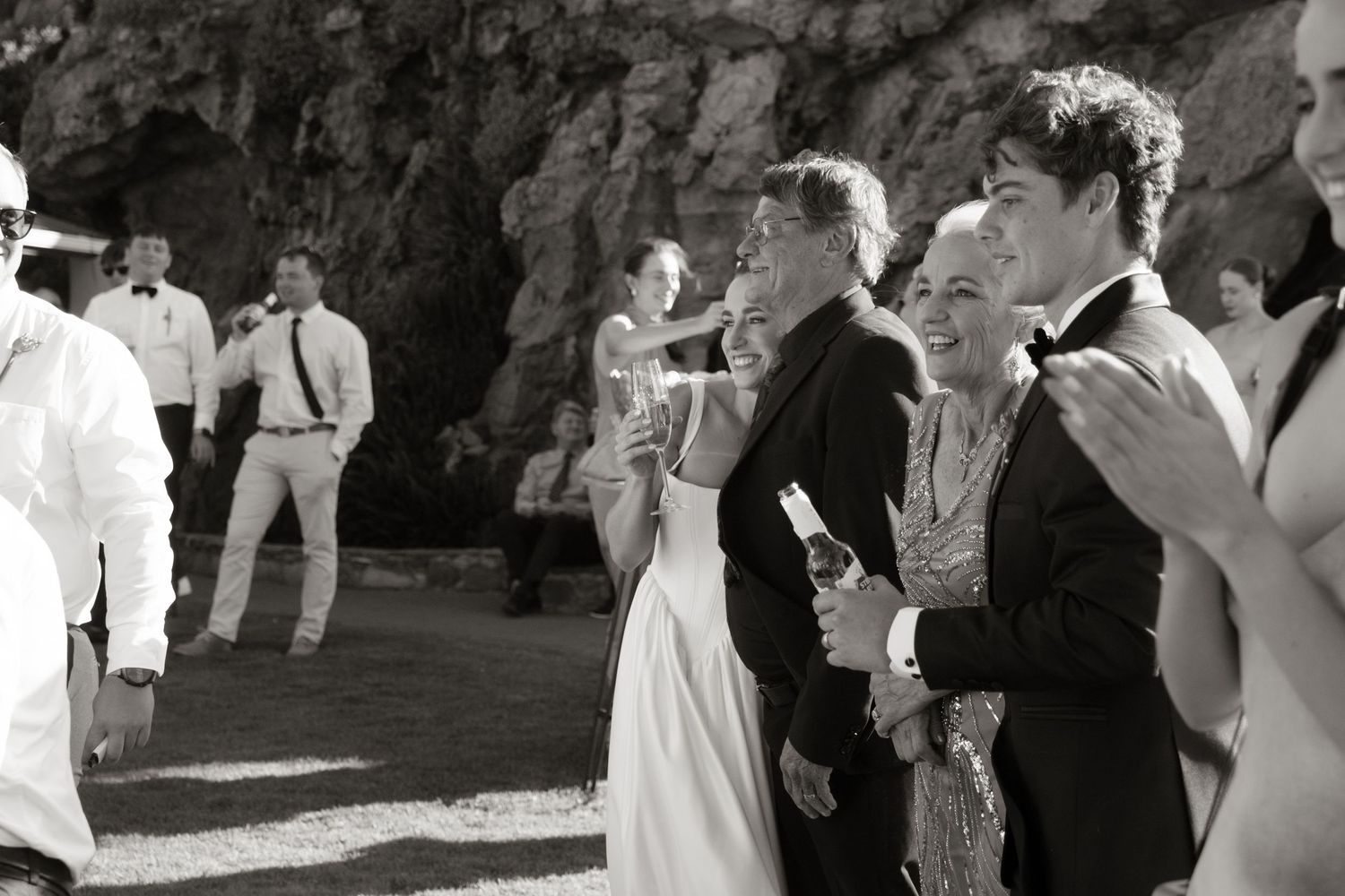 Wedding guests mingle and celebrate outdoors near rocky backdrop in classic black and white photography.
