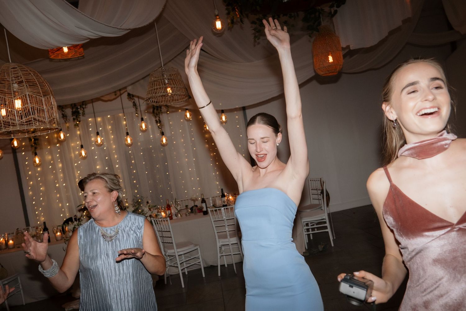 Three women dancing joyfully at a wedding reception under warm string lights and draped ceiling decor.