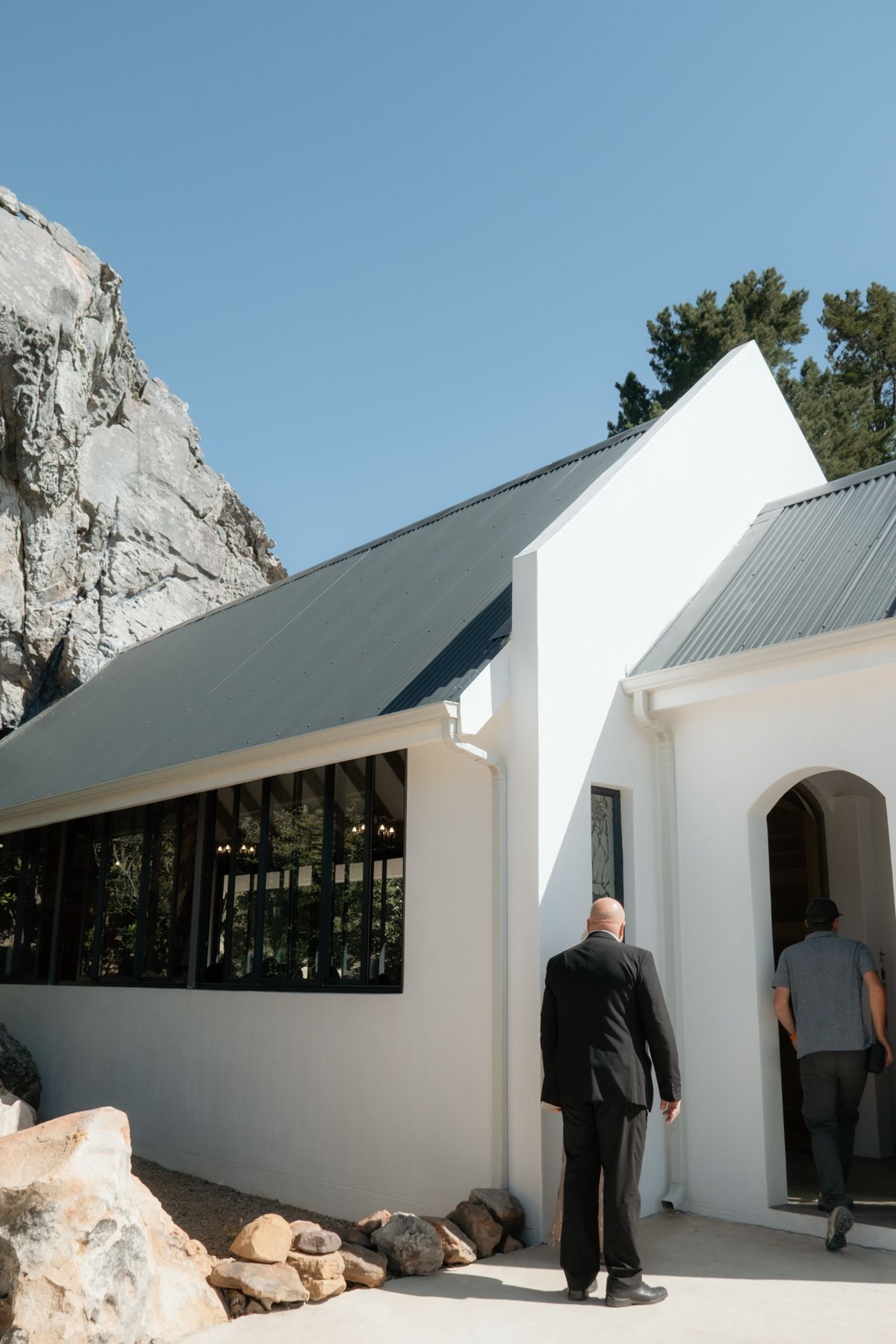 Two men in suits entering a white chapel nestled against dramatic rocky cliffs under a clear blue sky.