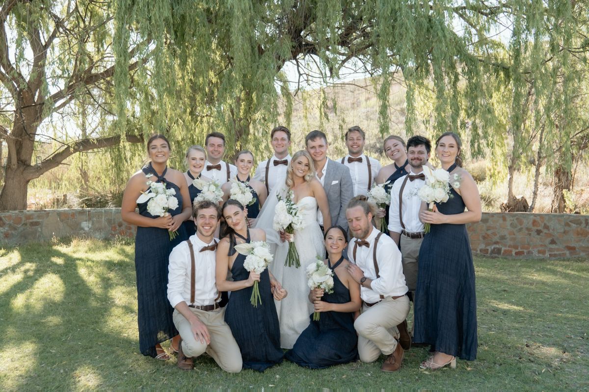 Wedding party poses under a willow tree, bridesmaids in navy dresses with groomsmen in suspenders and bow ties.