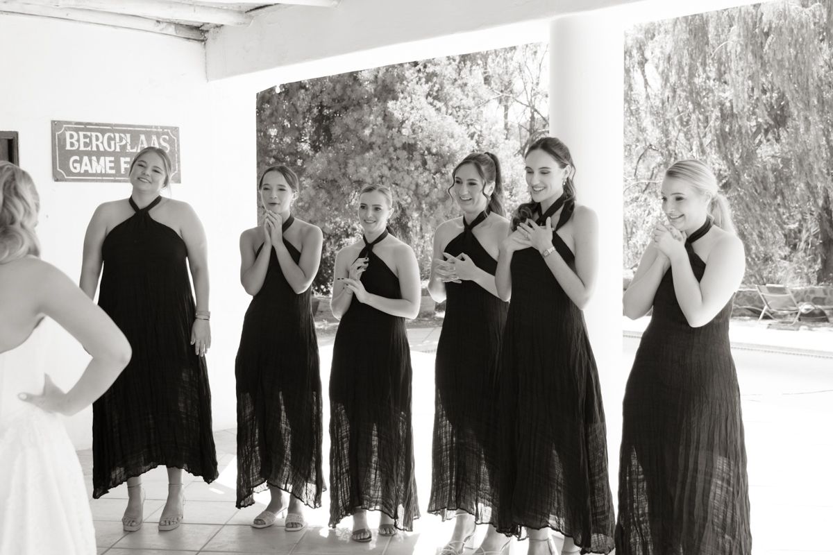 Black and white photo of bridesmaids in black dresses reacting emotionally to bride at outdoor wedding.