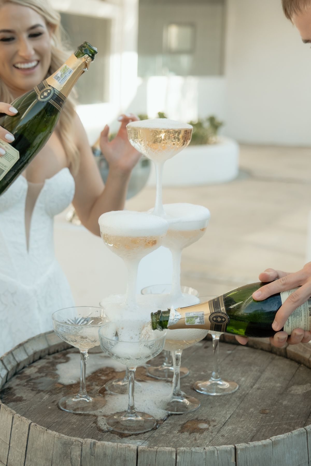 Bride laughing as champagne tower is poured on a rustic wooden barrel at wedding reception.