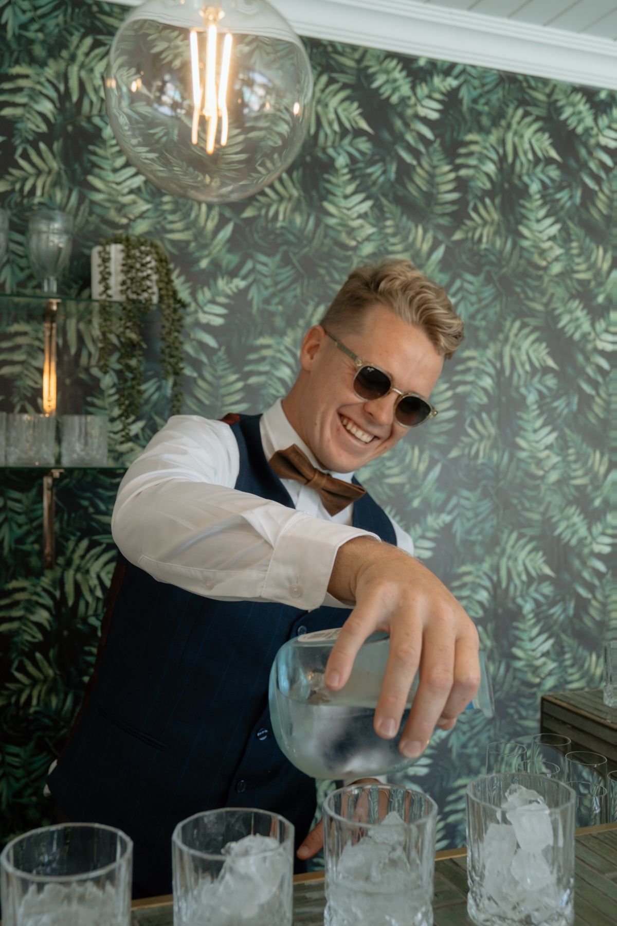 Smiling bartender in vest and sunglasses pours ice into glasses at wedding reception bar.