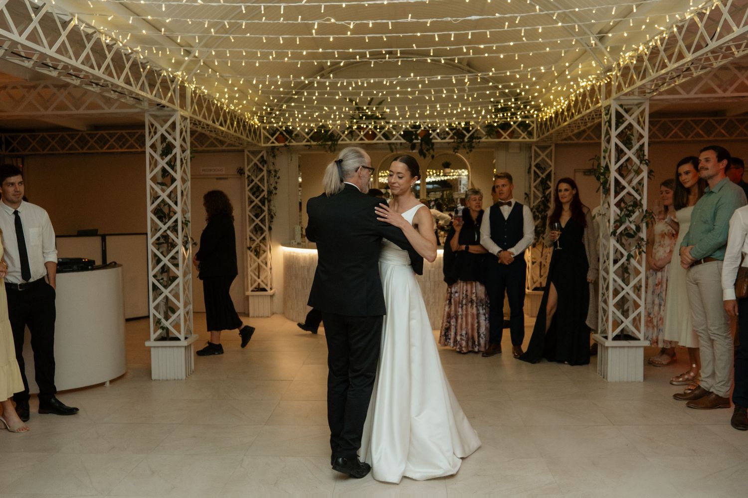 Bride and older gentleman share a father-daughter dance under fairy lights at wedding reception.