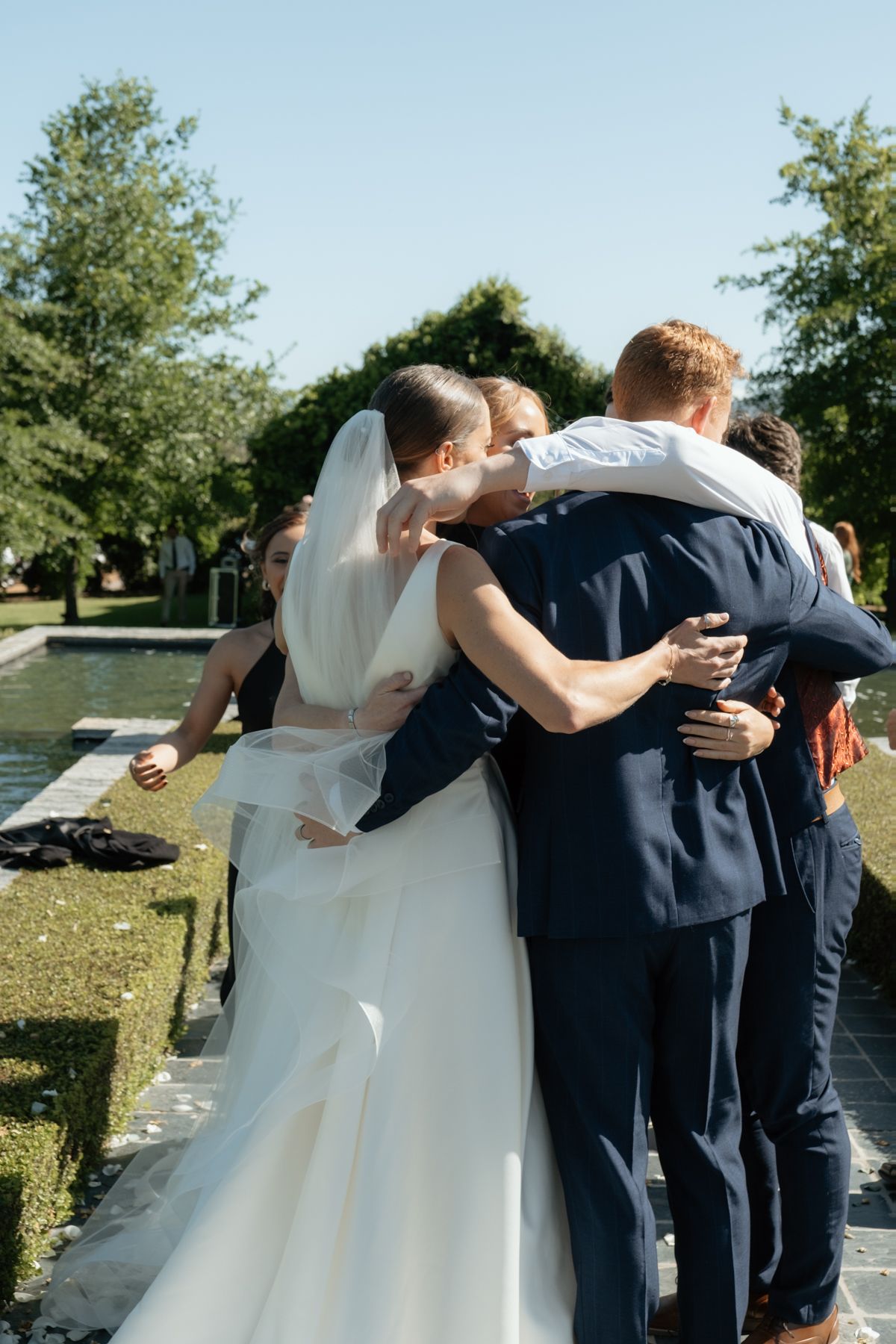 Bride in white gown and groom in navy suit share group hug with wedding party in sunny outdoor garden.