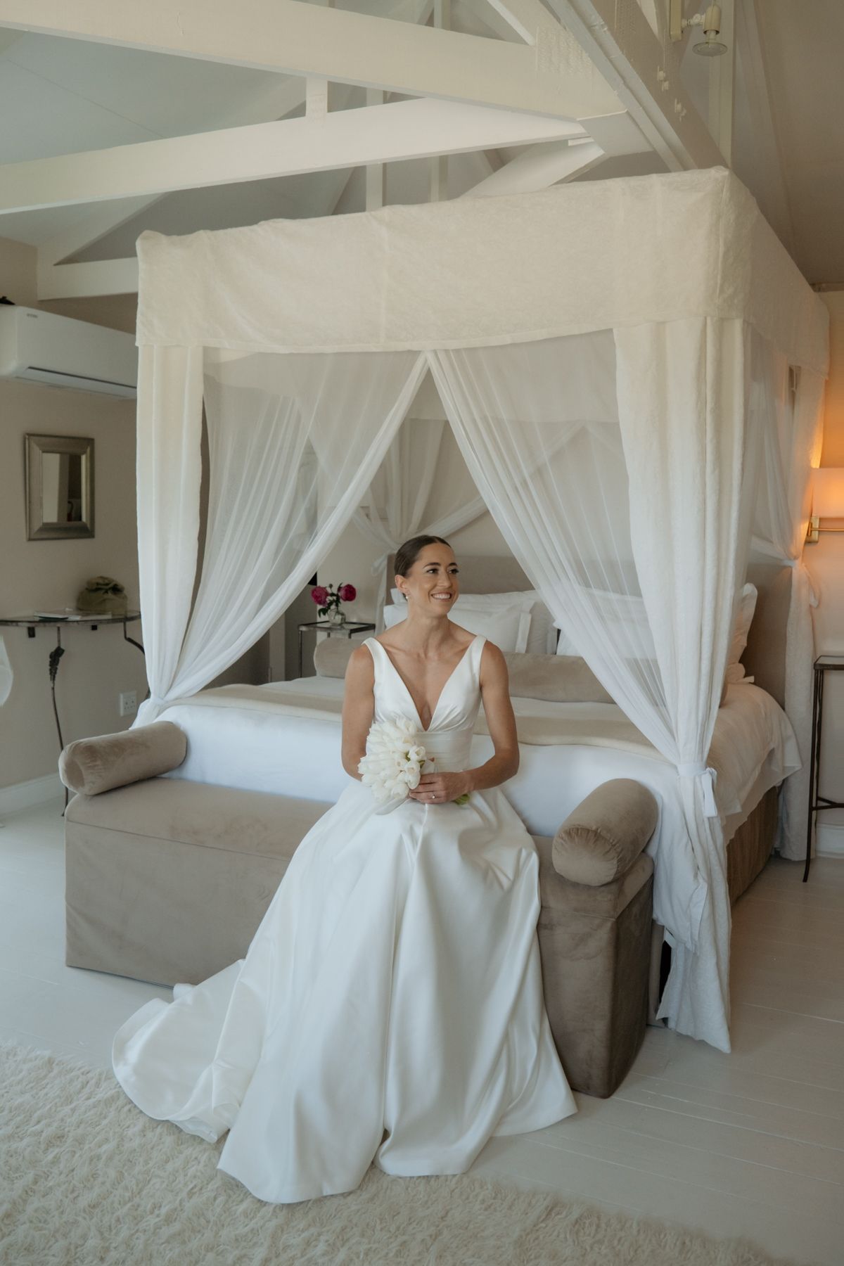 Elegant bride in white gown sits before a canopy bed holding a bouquet in a bright bridal suite.
