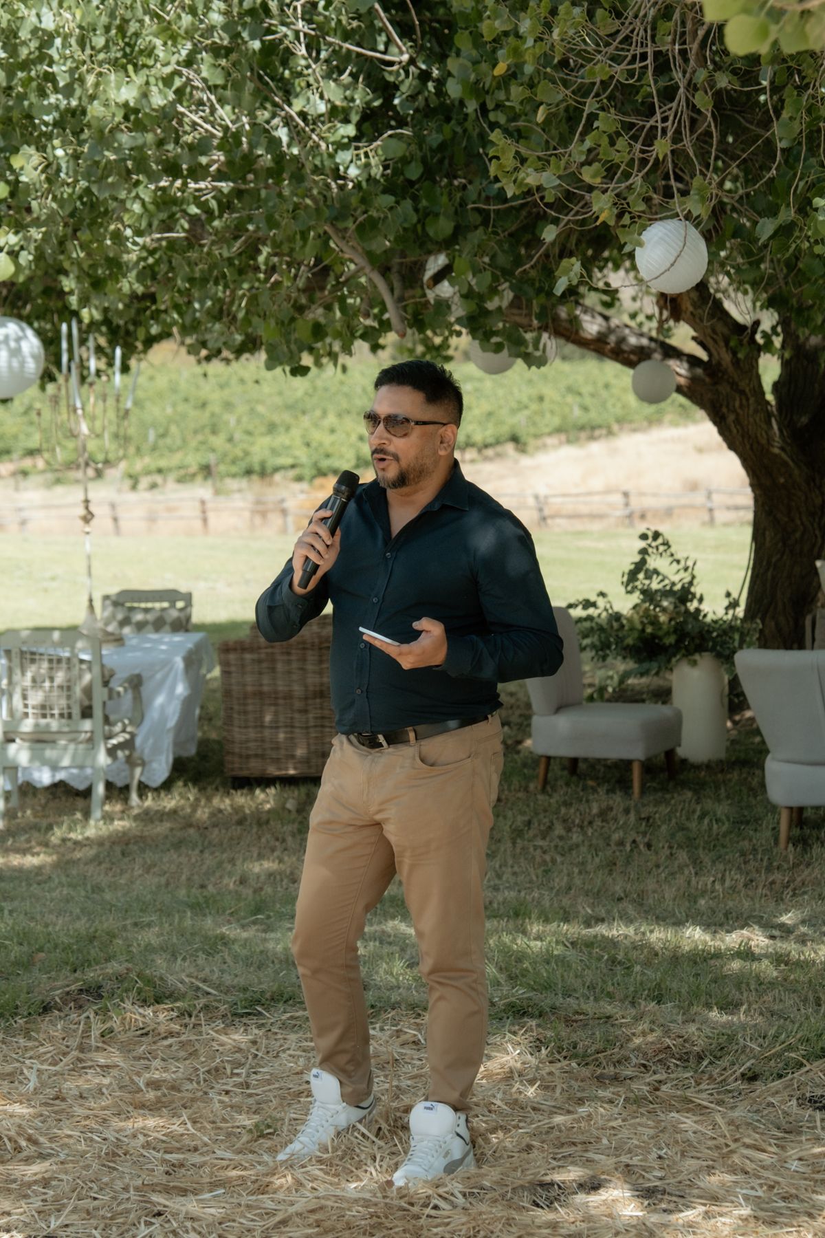 Man in dark shirt and khaki pants speaking into microphone at outdoor garden wedding reception.