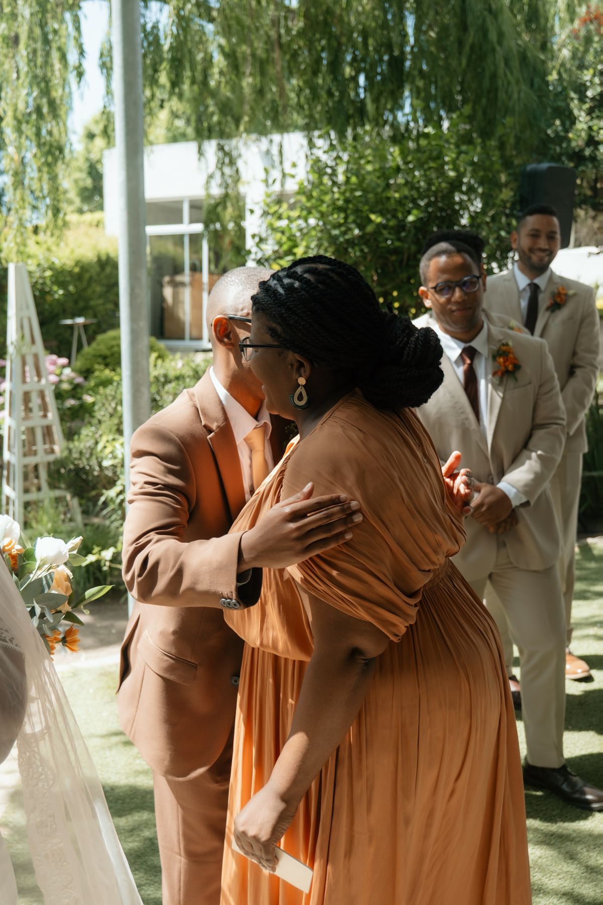 Elegant wedding guests embrace during an outdoor ceremony with groomsmen standing nearby amid lush greenery.