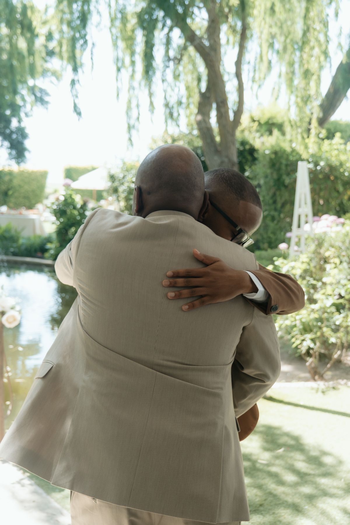 Two men in beige suits share a heartfelt embrace at an outdoor garden wedding ceremony.