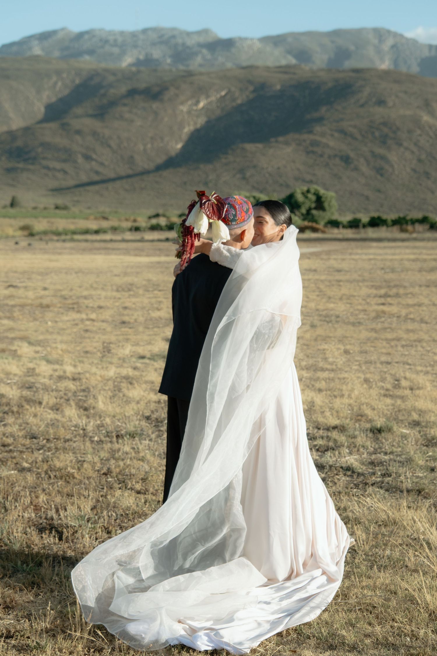 Bride and groom share romantic kiss in open field with stunning mountain backdrop on wedding day.